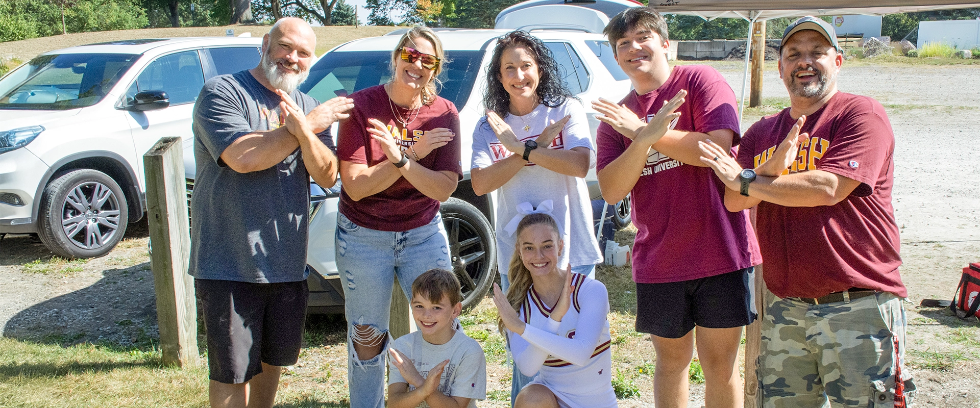 Group of Walsh University fans and a cheerleader smiling outdoors, posing with arms crossed in the swords up gesture.
