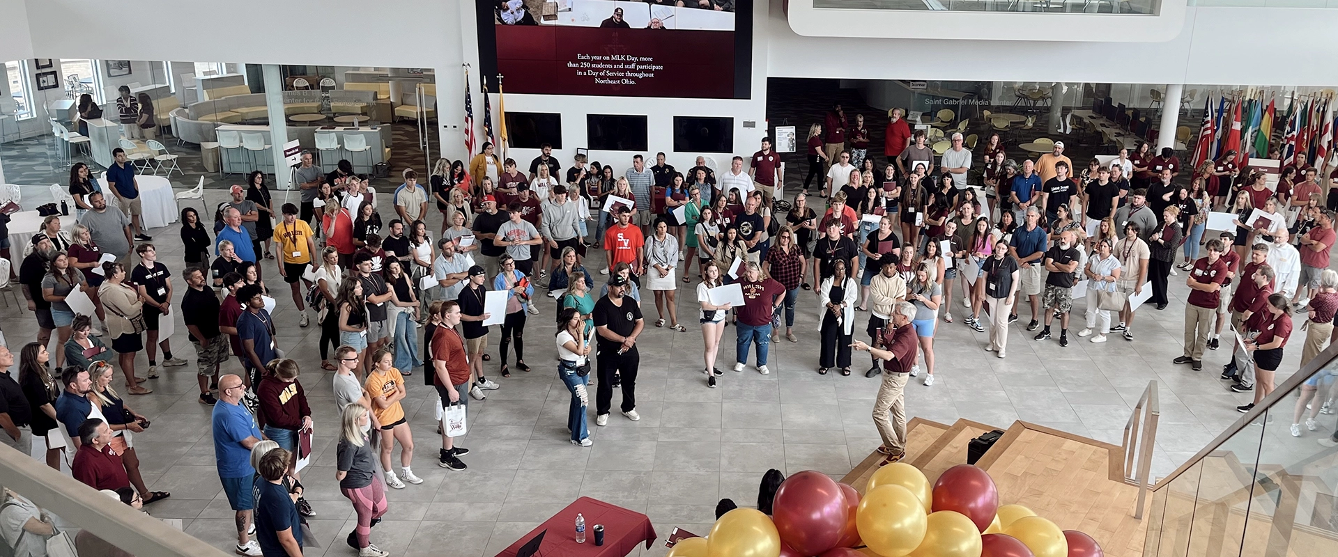 A large group of students stand inside a modern building listening to a man speak.