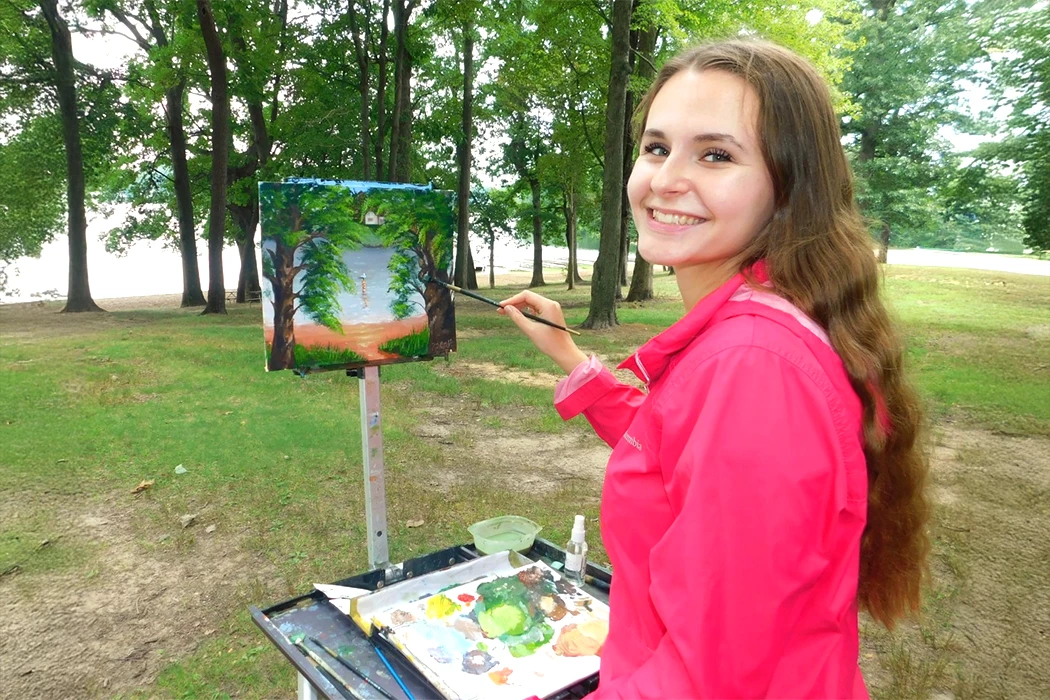 A young woman painting at an easel in a forested area near a lake.