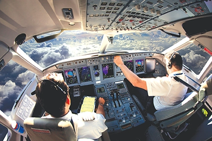 Cockpit view of pilots operating an aircraft, showcasing control panels and flight instruments above the clouds.