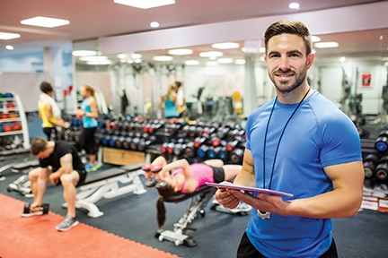 Fitness trainer smiling while holding a clipboard in a gym filled with various exercise equipment and people working out.