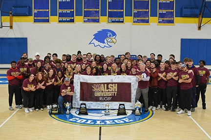 A group photo of the Walsh University rugby team celebrating with trophies, wearing school colors in a gymnasium setting.