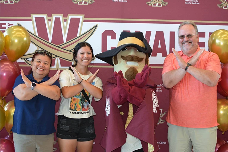 A new student and her family pose with the Walsh University mascot, Sir Walter, in front of a maroon backdrop.