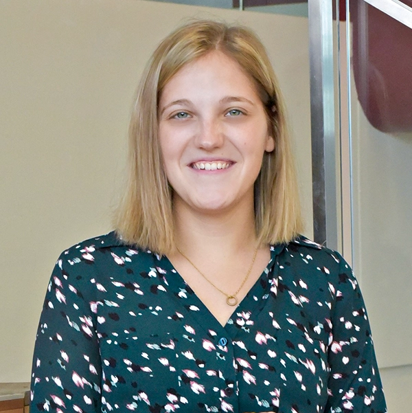 Portrait of a smiling woman with blonde hair, wearing a black and green patterned blouse, standing in an indoor setting.