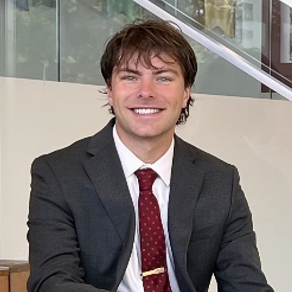 A smiling young man in a suit and tie, sitting in a modern office environment.