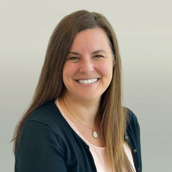 Professional headshot of a woman with long brown hair, wearing a black cardigan and a light pink top, smiling against a neutral background.