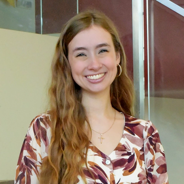 Smiling young woman with long wavy hair wearing a floral blouse, standing in a modern indoor space.