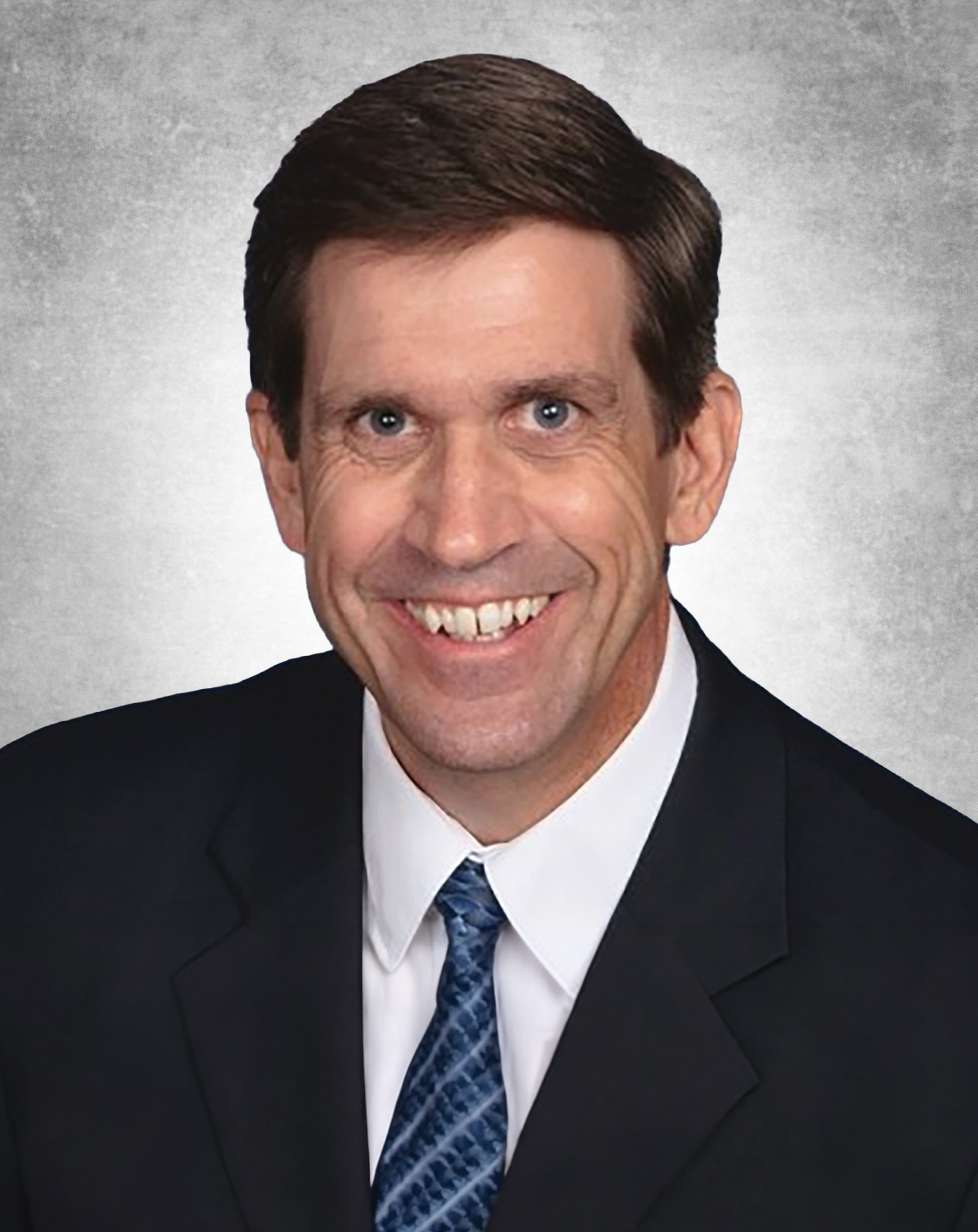 Studio headshot of Dr. James Monaghan, smiling in a dark suit and patterned blue tie against a gray background.