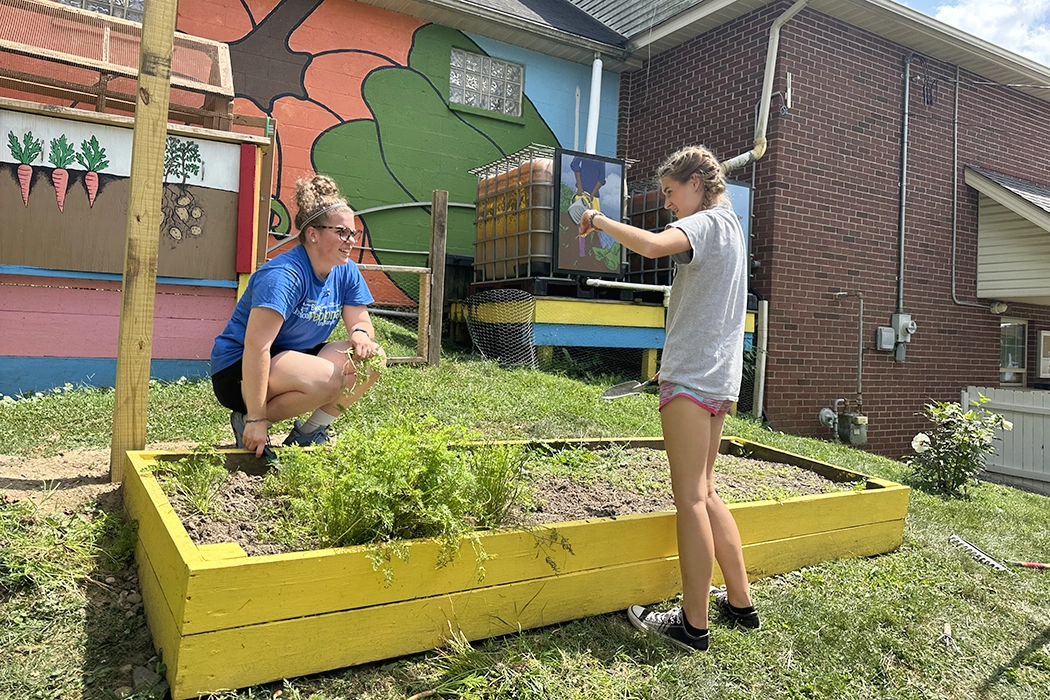 Two individuals tending to a community garden with vibrant painted backgrounds, one crouching and pulling weeds, while the other is pointing and guiding.