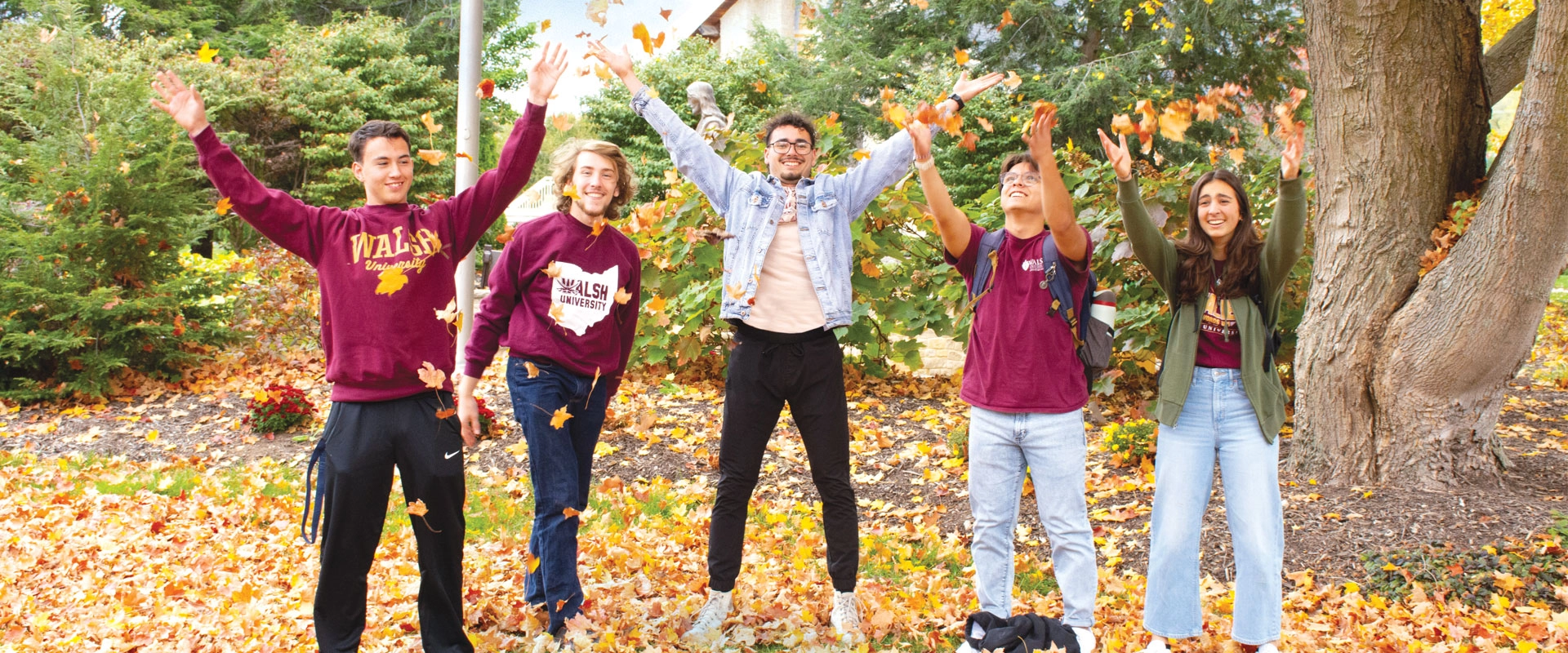 Four students outdoors in the Fall, tossing leaves in the air.