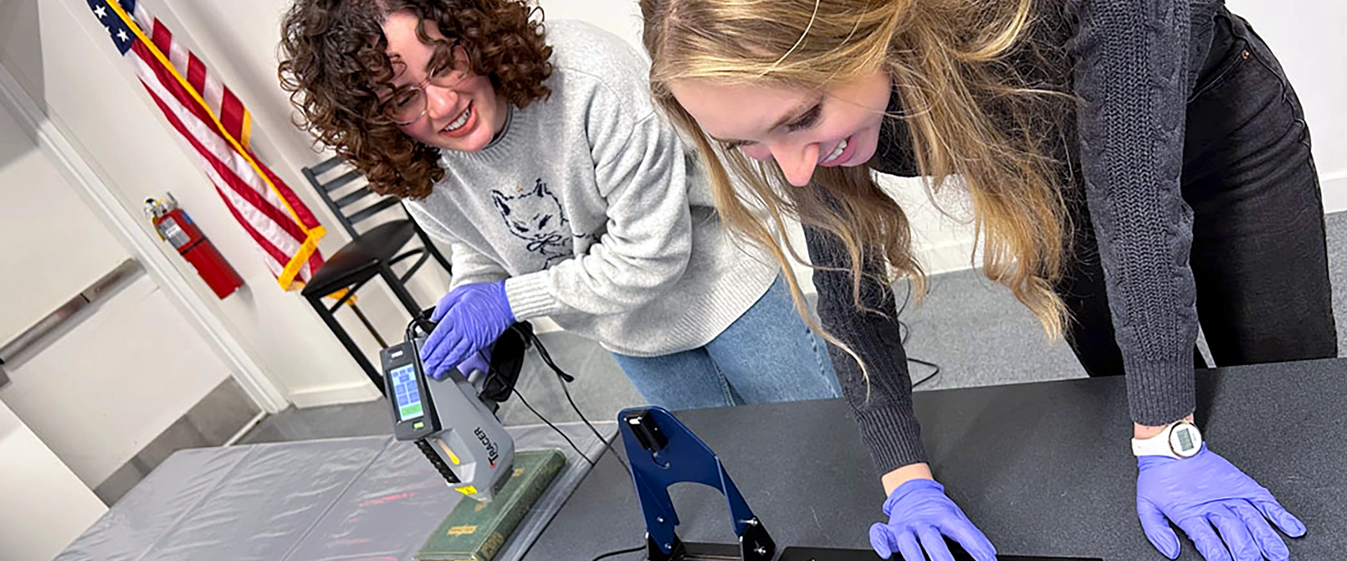 Two students wearing purple gloves work together in a lab setting, using a handheld X-ray fluorescence (XRF) analyzer to examine an artifact. They are smiling and engaged in the process.