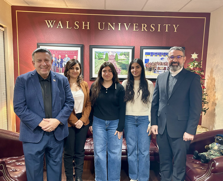 A group of four individuals, including a man in a blue blazer and three women in casual attire, pose together in front of a Walsh University sign and framed photos.