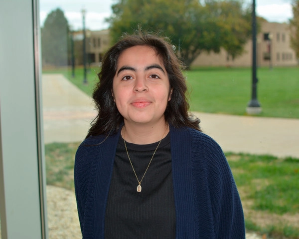 Young woman with shoulder-length dark hair, smiling while standing near a window with a green outdoor background.