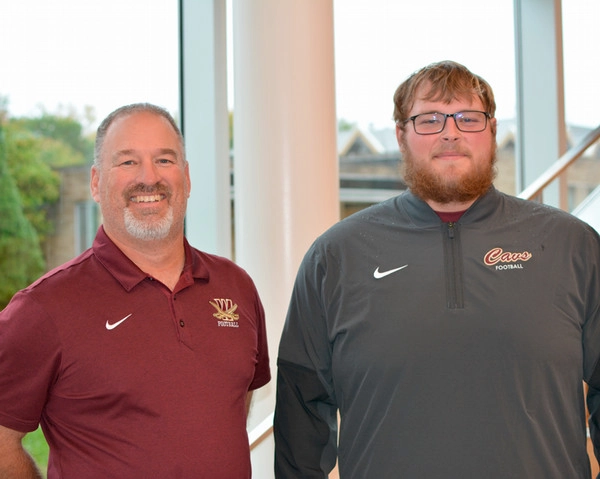 A football coach and his player stand side by side, smiling in a bright, indoor setting. They are wearing matching team apparel with logos.