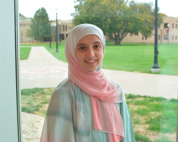 Smiling young woman in a light pastel outfit, standing indoors with a view of an outdoor campus space in the background.