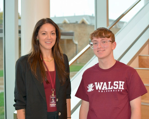 A woman in professional attire and a young man in a Walsh University t-shirt pose together near a staircase in a well-lit space.