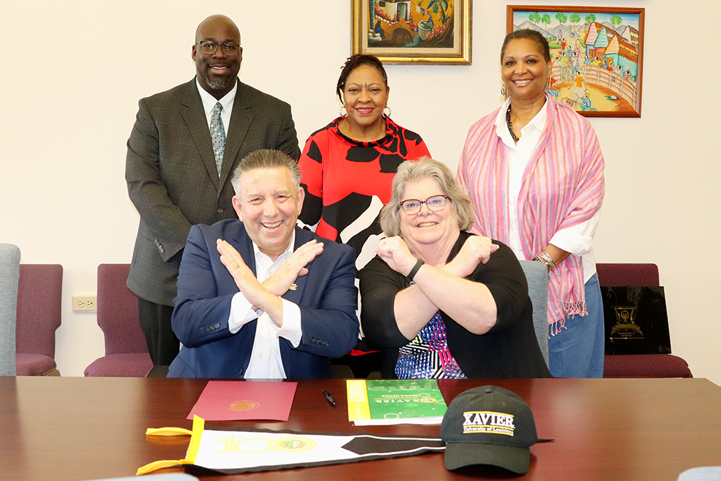 Five individuals, three standing and two seated at a table, pose for a photo to commemorate a partnership between Walsh University and Xavier University of Louisiana. Dr. Mike Dunphy and another seated individual cross their arms in a Swords Up! gesture.