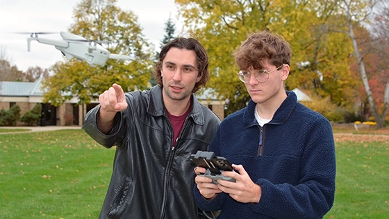 Photo of an instructor teaching a student how to fly a drone, with the drone hovering nearby and autumn trees in the background.